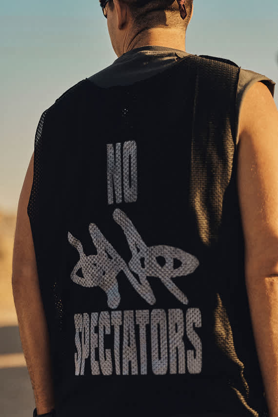 A close-up from behind a person wearing a black mesh vest that reads "NO SPECTATORS" in bold, textured lettering.