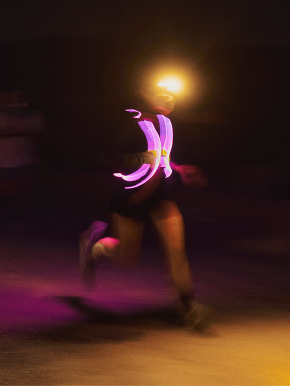 A blurry, long-exposure shot of a person running at night, illuminated by a glowing pink safety vest and a bright yellow headlamp.
