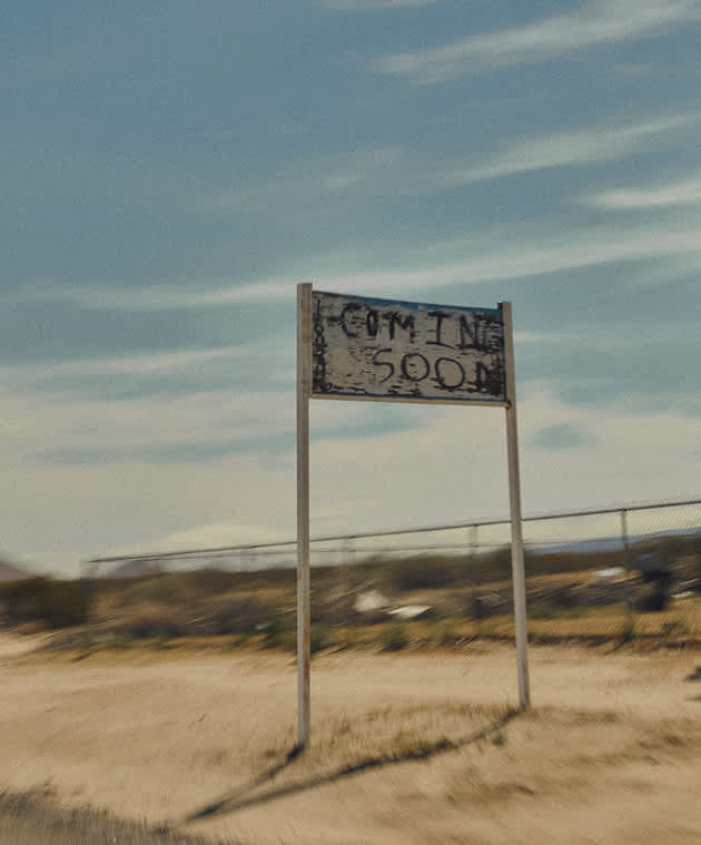 A weathered, white wooden sign that says "COMING SOON" standing in a sandy desert area under a cloudy sky.