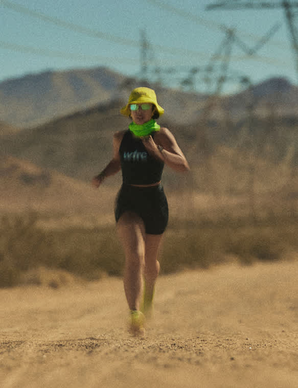 A runner wearing a yellow bucket hat, green neck gaiter, and sunglasses running down a dirt road in a desert landscape.