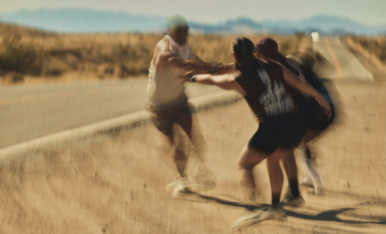 A blurry action shot of runners cheering and reaching out to a teammate on the side of a desert road.