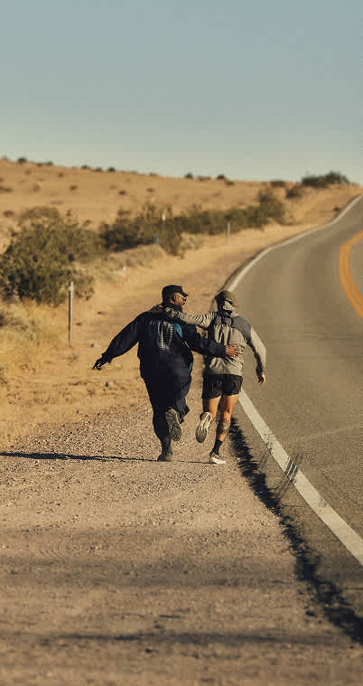 Two people running together down the shoulder of a paved desert highway under a clear sky.