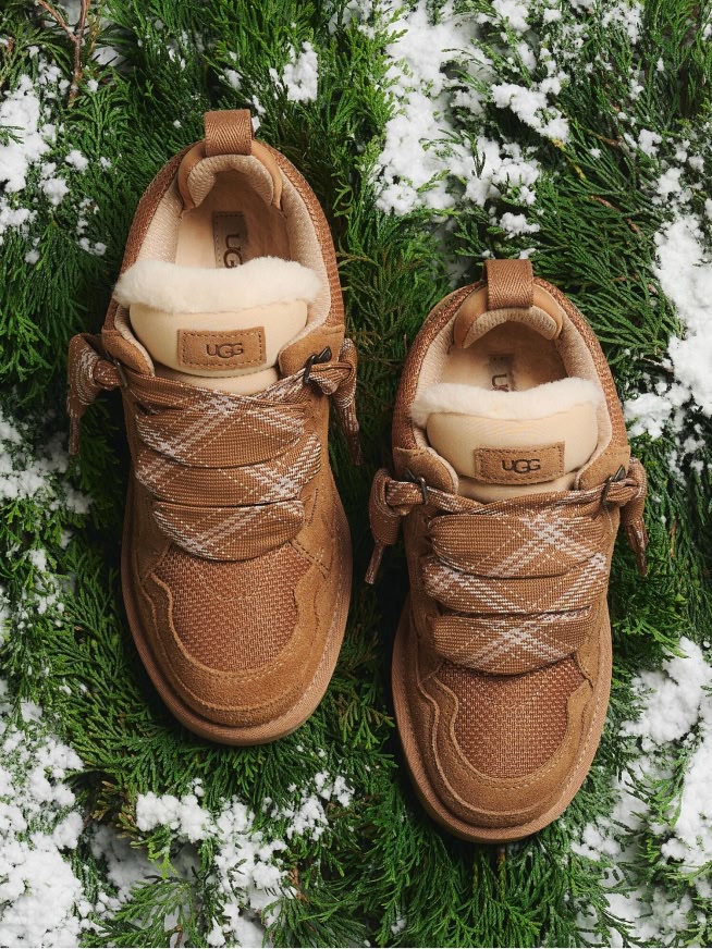 Two UGG sneakers, brown with plaid laces & fur trim, on snowy evergreen branches.