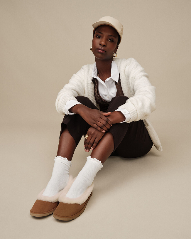 Person wearing white cardigan, white shirt, dark overalls and brown slippers with socks.