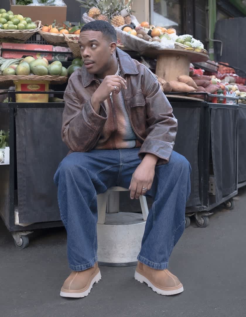 Marvin wearing a brown leather jacket and blue jeans with the Tasman Lug, sitting on a stool in front of a vegetable stand. Marvin standing in front of a Parisian supermarket, wearing blue jeans and a brown leather jacket with the Tasman Lug.