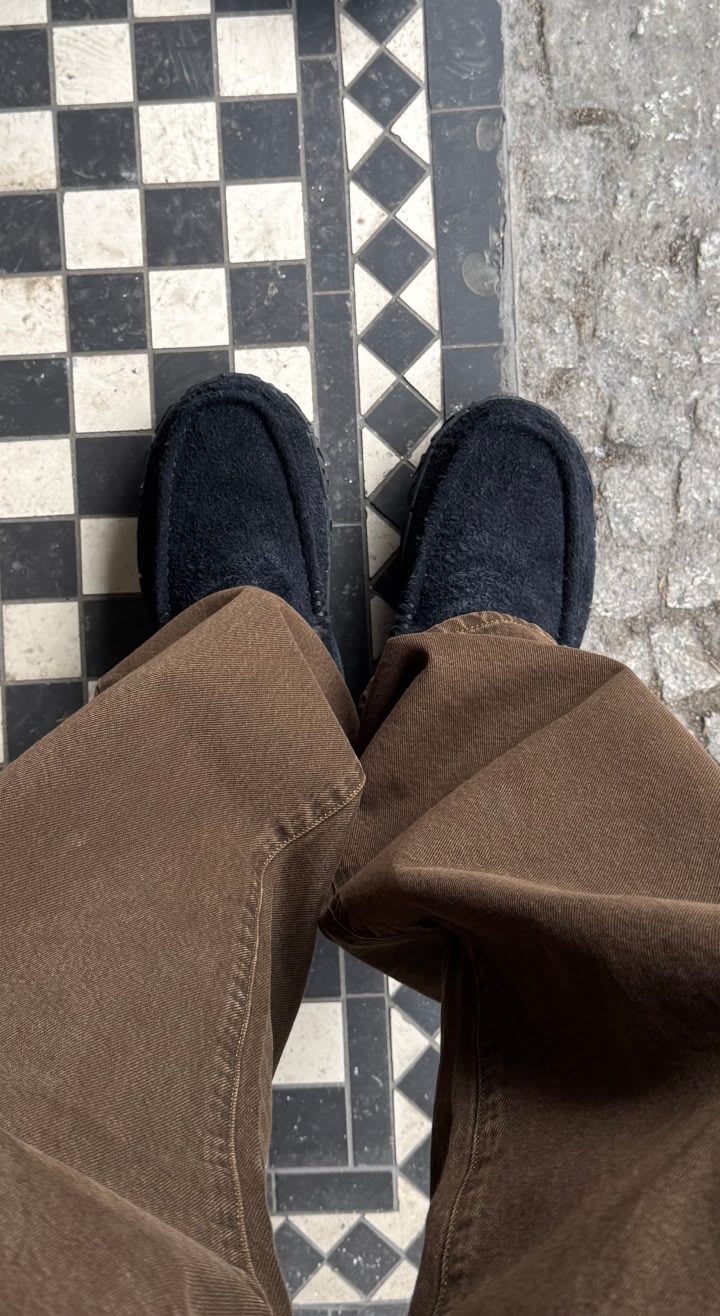 A downward shot of a person's legs wearing brown keans with the Ascot Vibram Wrap in Black on a chequered floor.