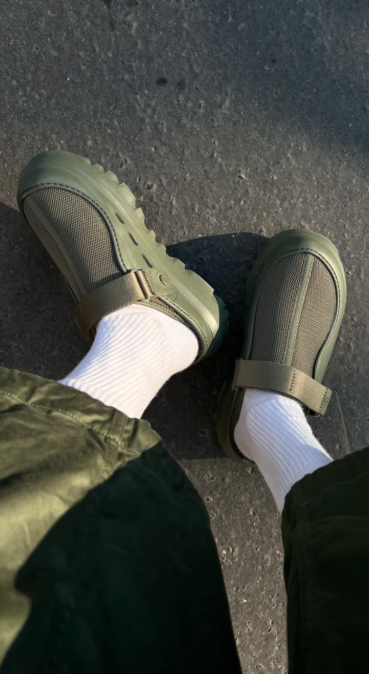 A downward shot of a person's legs wearing green trousers, white socks and the PeakMod in Woodland Green.