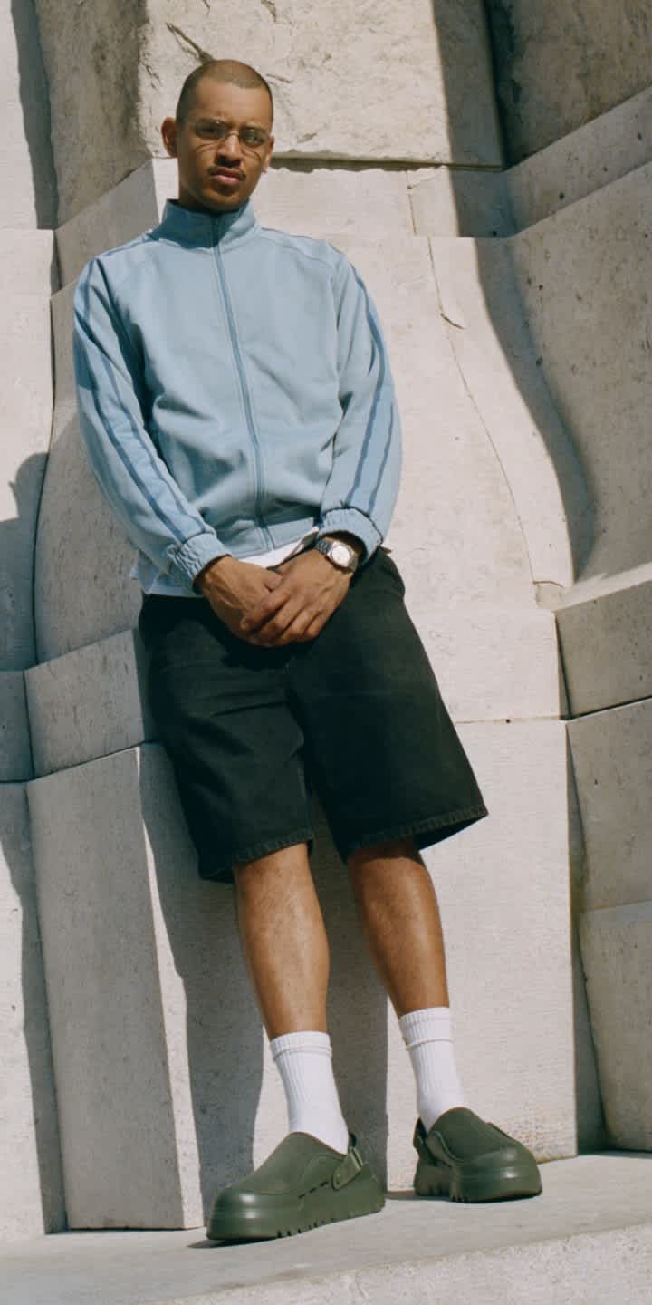 A person standing on a concrete ledge, wearing a light blue track jacket with shorts and the Peakmod in Woodland Green.