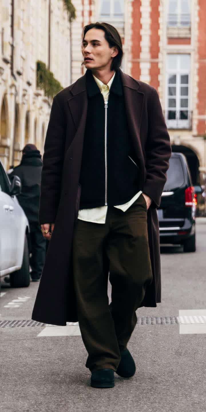 A person wearing a dark zip jacket with dark trousers and the Classic Mini Boot in Black walking down a road. 