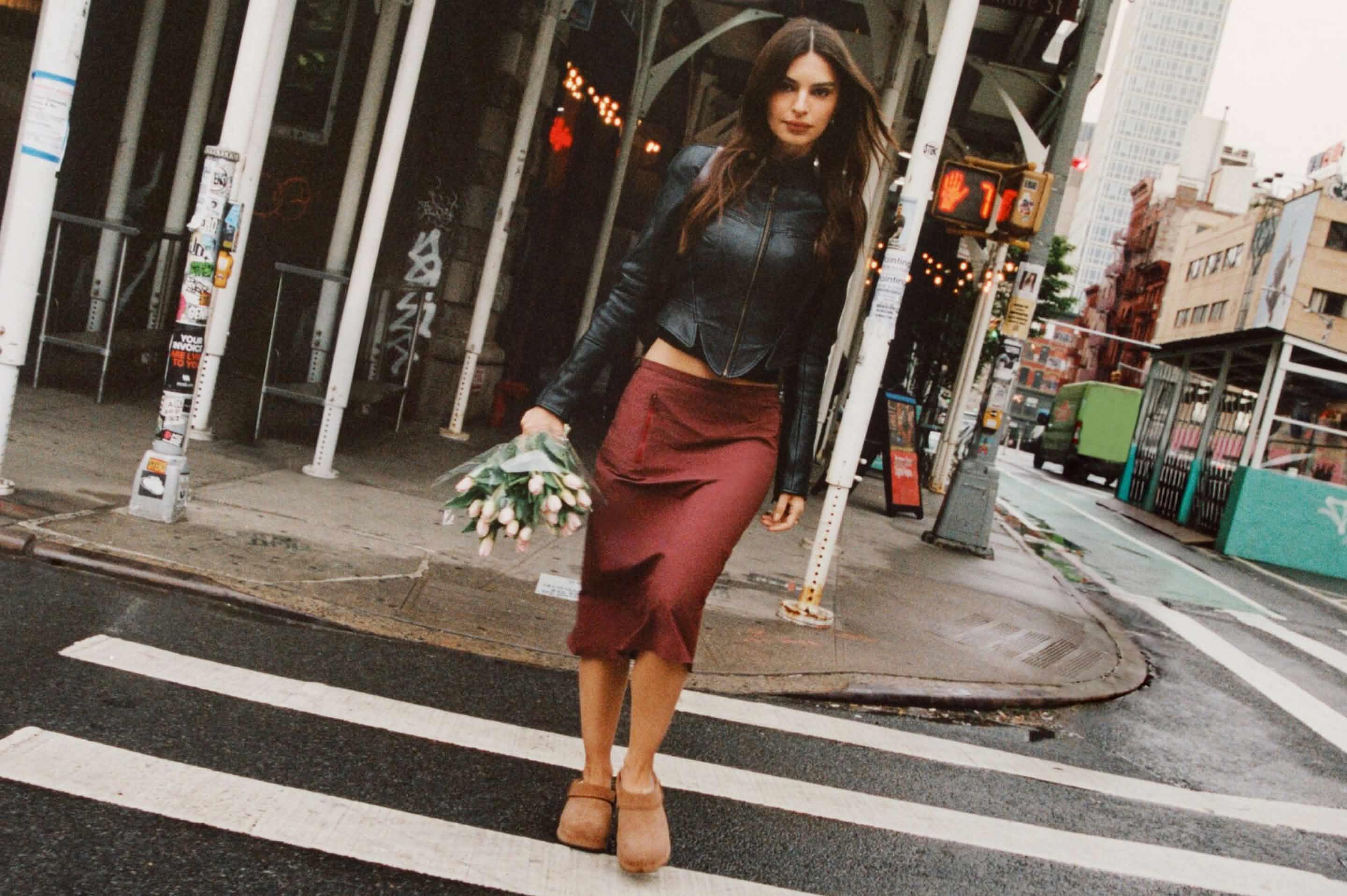 UGG brown suede clogs worn with a burgundy skirt on a city crosswalk.