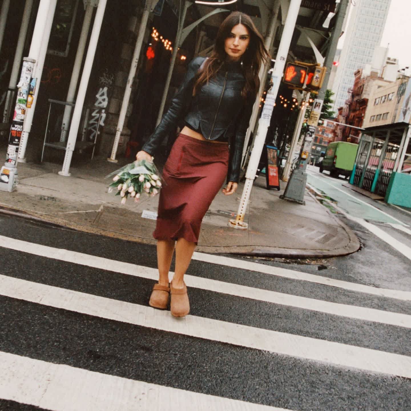 UGG brown suede clogs worn with a burgundy skirt on a city crosswalk.