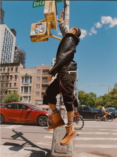  Person in leather jacket and tan boots balancing on a pillar, reaching up on a city