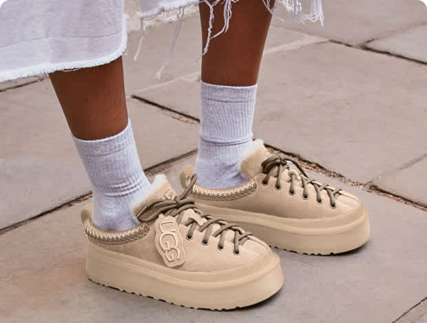Person wearing light beige UGG slippers with thick soles and socks on a paved