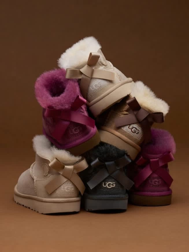 Stacked UGG mini boots with bows in cream, pink, gray on a brown background.