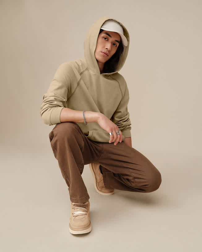 Model crouching in a beige hoodie, brown trousers and suede lace-up shoes.