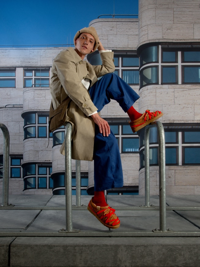 Person in beige beret, trench coat, blue trousers, red socks, tan shoes on railing.