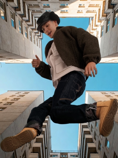 Person in hat, jacket, jeans, boots jumping high between tall buildings, blue sky