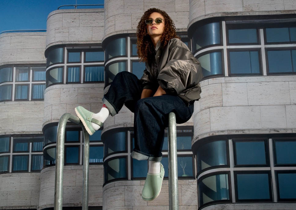 Person in bomber jacket, jeans, sunglasses, and green clogs sitting on a rail.