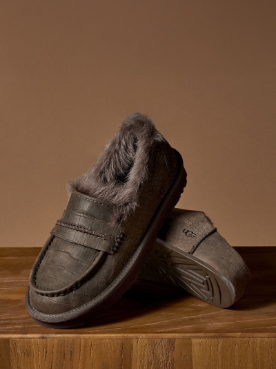Pair of brown crocodile-textured UGG slippers with fur lining on wood surface.